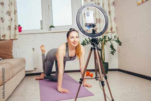 A young woman video blogger is livestreaming her workout from home using a smartphone and a ring light mounted on a tripod