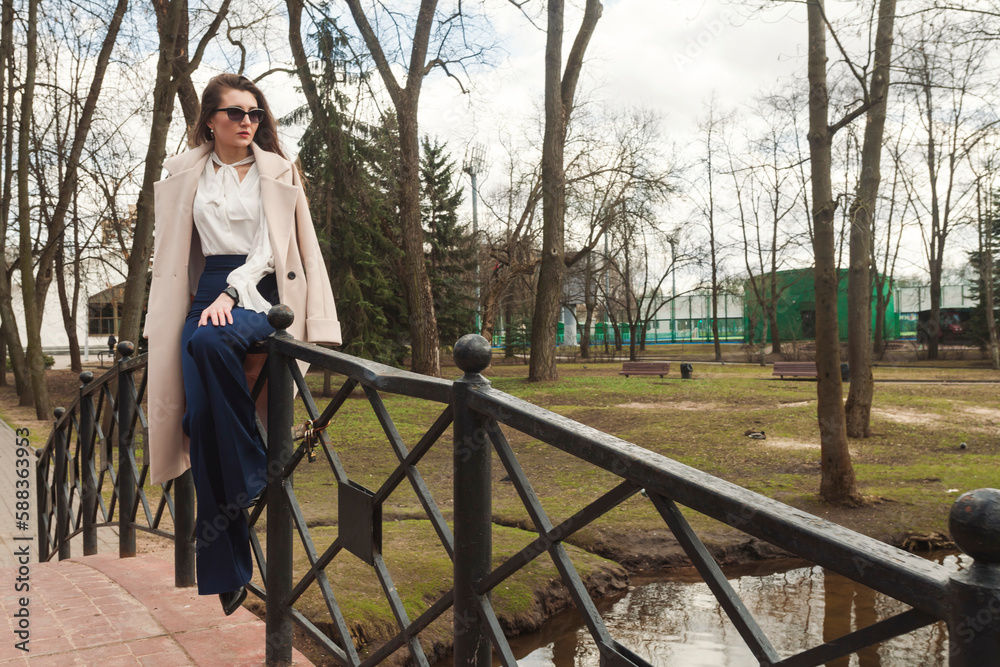 Fashionable girl sitting on the rails of a bridge over a creek in the park, with a coat thrown over her shoulders, smart watch on her wrist, stylish tight dark blue pants, white shirt.