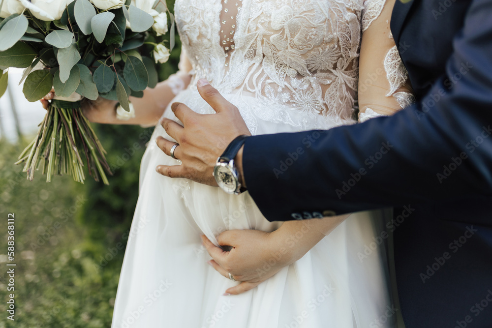 A young couple just married carrying their first child. Delicate wedding bouquet in the hands of the bride. Groom hugging bride from behind.