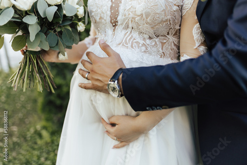 Obraz na płótnie A young couple just married carrying their first child. Delicate wedding bouquet in the hands of the bride. Groom hugging bride from behind. w salonie