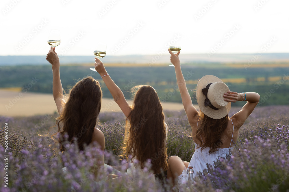 Three friends, young women with wine glasses in hands sitting in lavender flowers. Picnic on the lavender field.