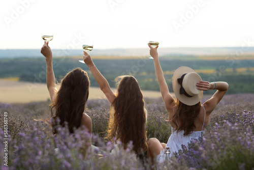 Obraz na płótnie Three friends, young women with wine glasses in hands sitting in lavender flowers. Picnic on the lavender field. w salonie