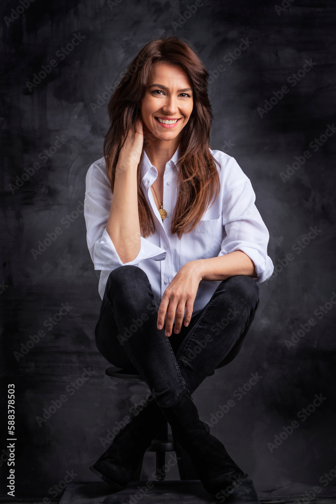 Studio portrait of a brunette haired attractive woman wearing white shirt and black jeans while posing at isolated dark background Studio portrait of a brunette haired attractive woman wearing white shirt and black jeans while posing at isolated dark background