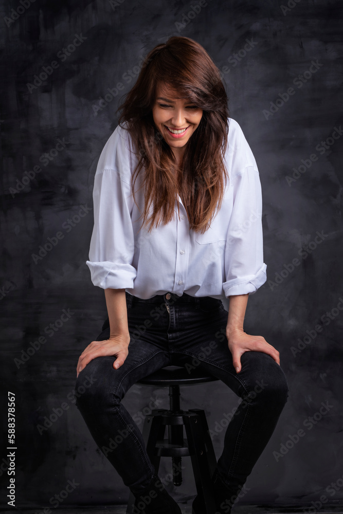 Naklejka premium Studio portrait of a brunette haired attractive woman wearing white shirt and black jeans while posing at isolated dark background