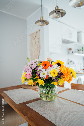 Kitchen counter table with focus on vase with huge multicolor various flower bouquet with blurred background of modern cozy white kitchen. Home interior design details. Mother's day. Copy space.