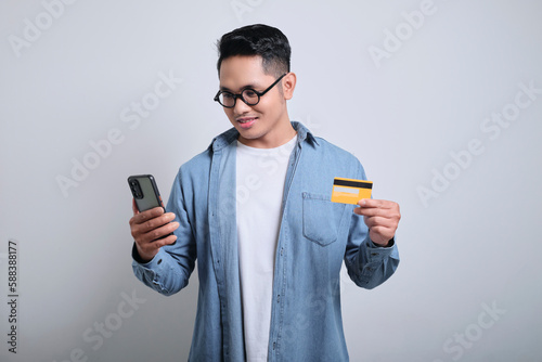 Smiling young asian man wears denim shirt using mobile cell phone hold credit bank card doing online shopping order delivery