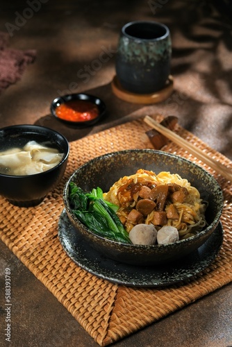 A bowl of Chicken noodles in rustic bowl. Meat ball, chilli sauce in the back. Chopstick beside the bowl. Mie ayam photo