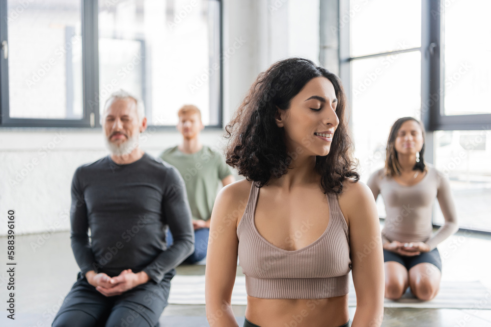 Smiling middle eastern woman meditating near blurred people in yoga class.