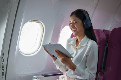 Young Asian attractive business woman passenger sitting on business class luxury plane while wearing wireless headphone for relax and working with tablet during the flight