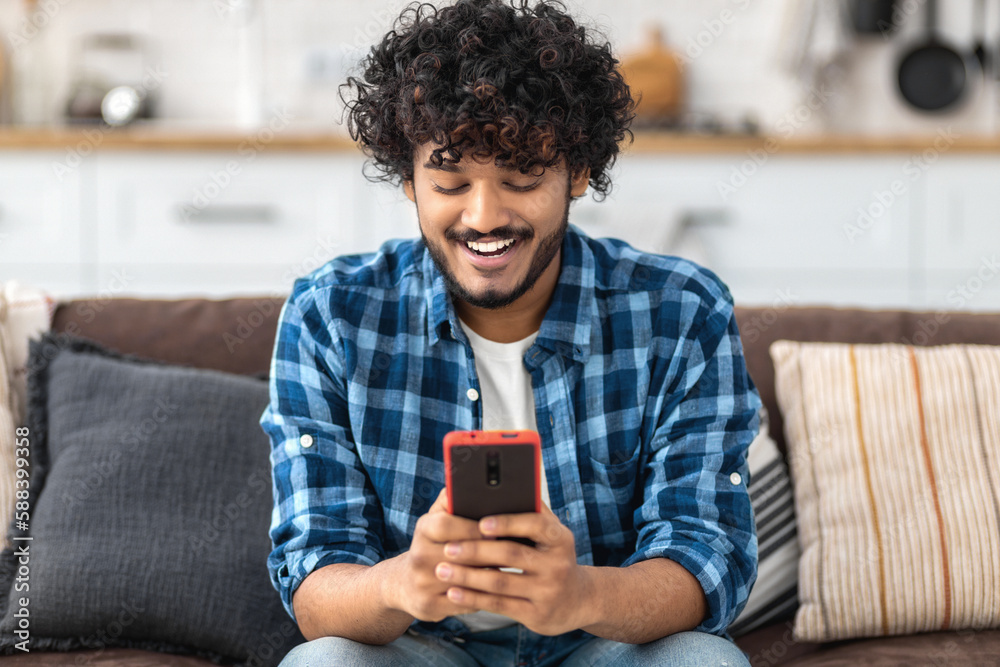 Happy Indian man hanging on social media using mobile phone. Smiling Asian male shopping online and payment sitting at home on the sofa Happy Indian man hanging on social media using mobile phone. Smiling Asian male shopping online and payment sitting at home on the sofa