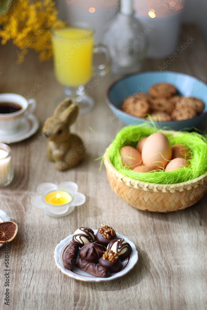 Easter eggs in the basket, bowl of cookies, chocolate pralines, Easter bunny figurine, cups of tea, glasses of juice, flowers and lit candles on the table. Selective focus. Easter eggs in the basket, bowl of cookies, chocolate pralines, Easter bunny figurine, cups of tea, glasses of juice, flowers and lit candles on the table. Selective focus.