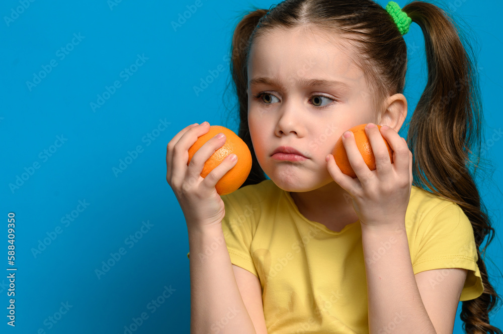 A small and smiling girl is holding two tangerines on a blue background. A small and smiling girl is holding two tangerines on a blue background.