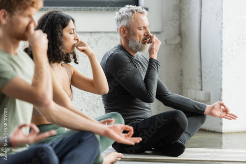 Mature man practicing nostril breathing and gyan mudra near interracial people in yoga studio. photo