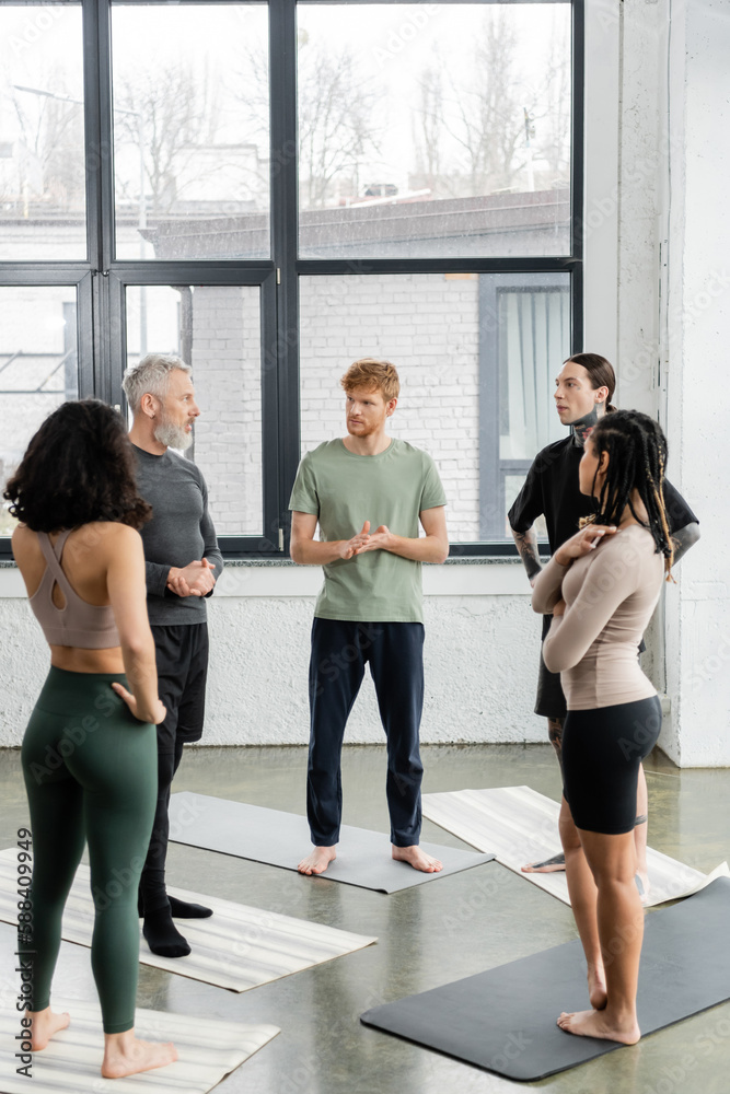 Interracial people looking at mature coach while standing on mats in yoga class.