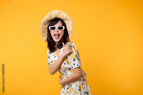 Portrait beautiful young asian woman wearing springtime dress and pointing to copy space for advertising isolated on yellow background. Summertime advertising banner concept.