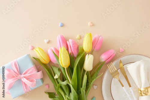 Fototapeta Naklejka Na Ścianę i Meble -  Mother's Day concept. Top view photo of plate with cutlery and fabric napkin gift box and bouquet of flowers yellow pink tulips and colorful hearts on isolated pastel beige background