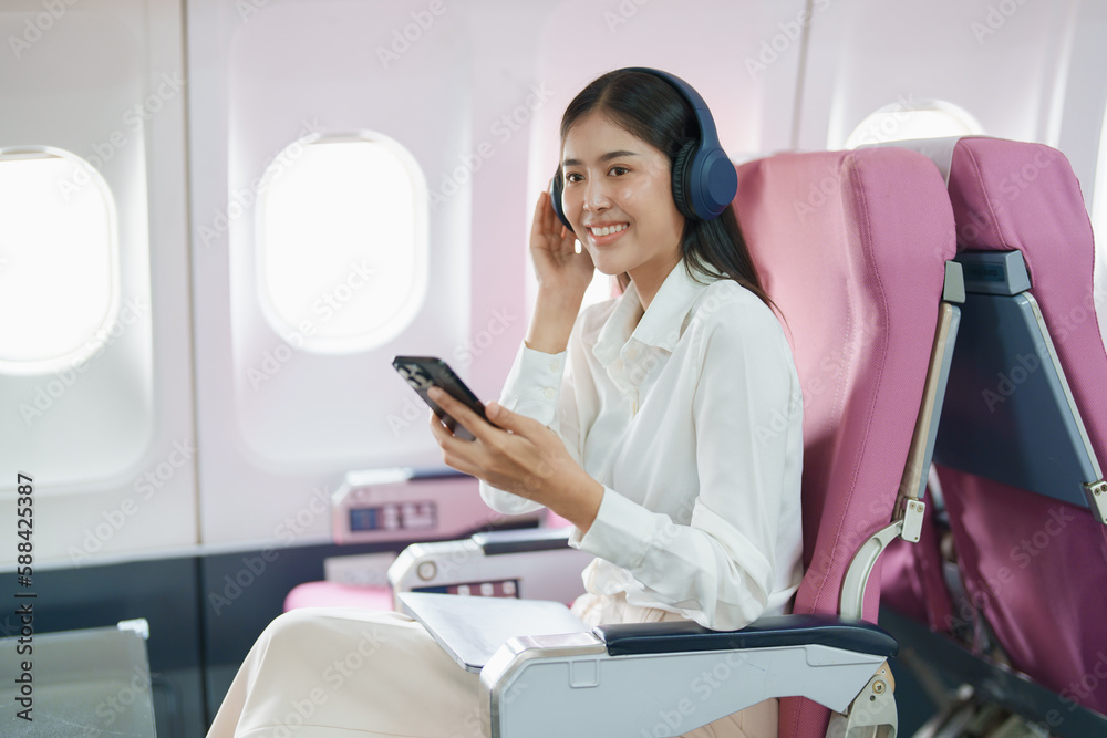 Young Asian attractive business woman passenger sitting on business class luxury plane while wearing wireless headphone and using smart phone mobile for relax during the flight Young Asian attractive business woman passenger sitting on business class luxury plane while wearing wireless headphone and using smart phone mobile for relax during the flight