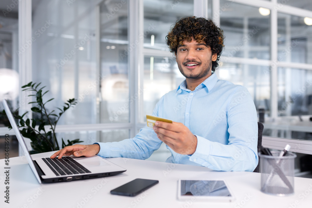 A young Muslim man is sitting in the office, working on a laptop and using a credit card. A young Muslim man is sitting in the office, working on a laptop and using a credit card.