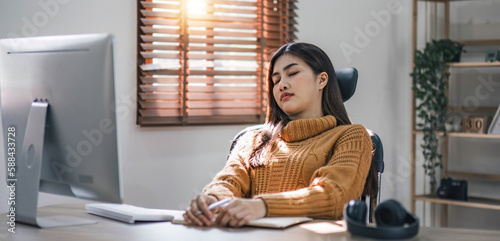 Tired teen asian lady in headphones falls asleep at table with laptop in room interior. Boring video lesson at home, problems at study, education with modern technology, online webinar and overwork © wichayada