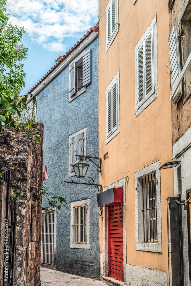 Fototapeta premium Buildings with bright blue and yellow facades on a narrow street in the Old Town of Budva, Montenegro. Houses with wooden shuttered windows and an old glass street lamp in Stari Grad, vertical