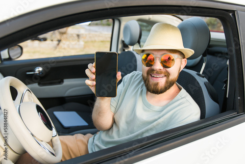Smiling bearded man in sunglasses showing mobile phone with empty screen for mock up sitting on driver seat in new luxury car cabriolet  holding gadget in hand  selective focus on device