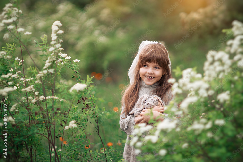 A cute little girl with brown hair holds favorite fluffy toy bunny among white flowers in the park. Background summer green flowering on the lawn. Child playing on the spring nature outdoors. Soft A cute little girl with brown hair holds favorite fluffy toy bunny among white flowers in the park. Background summer green flowering on the lawn. Child playing on the spring nature outdoors. Soft