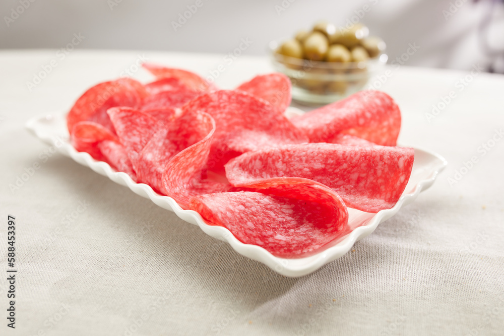 salami slices on a white plate with olives in the background. sausage close-up. meat delicacy on a sunny day. traditional mediterranean snack on a light surface. dried sausage serving. antipasta macro salami slices on a white plate with olives in the background. sausage close-up. meat delicacy on a sunny day. traditional mediterranean snack on a light surface. dried sausage serving. antipasta macro
