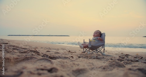 happy cute adorable Asian baby infant sitting relaxing on little chair and smiling with waves on background at seaside tropical sandy beach in sunset sunrise during holiday vacation summertime