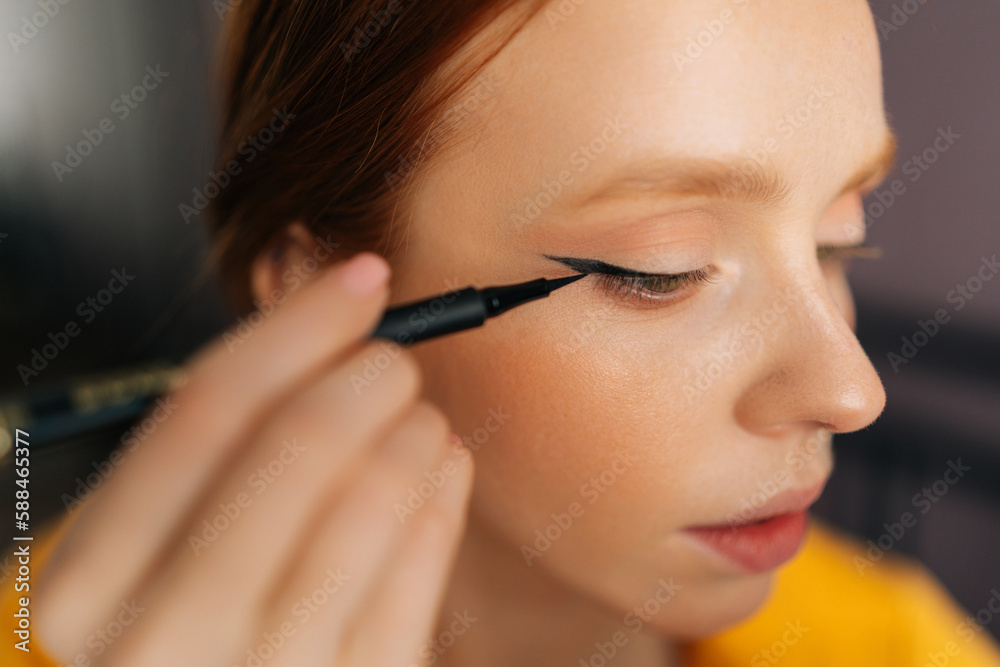 Closeup face of red-haired young woman using contour brush drawing arrows on eyelids looking at reflection in mirror. Beautiful redhead female applying eyeliner drawing cat eyes makeup sitting on bed.