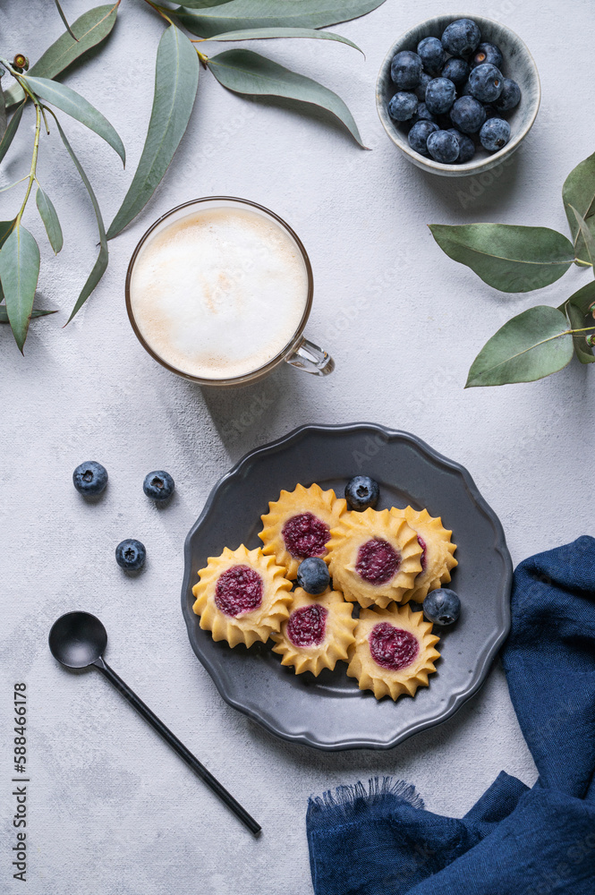Cappuccino or latte with milk foam in a cup with homemade berry cookies in a dark plate and blueberries on a light background with eucalyptus branches. Cappuccino or latte with milk foam in a cup with homemade berry cookies in a dark plate and blueberries on a light background with eucalyptus branches.