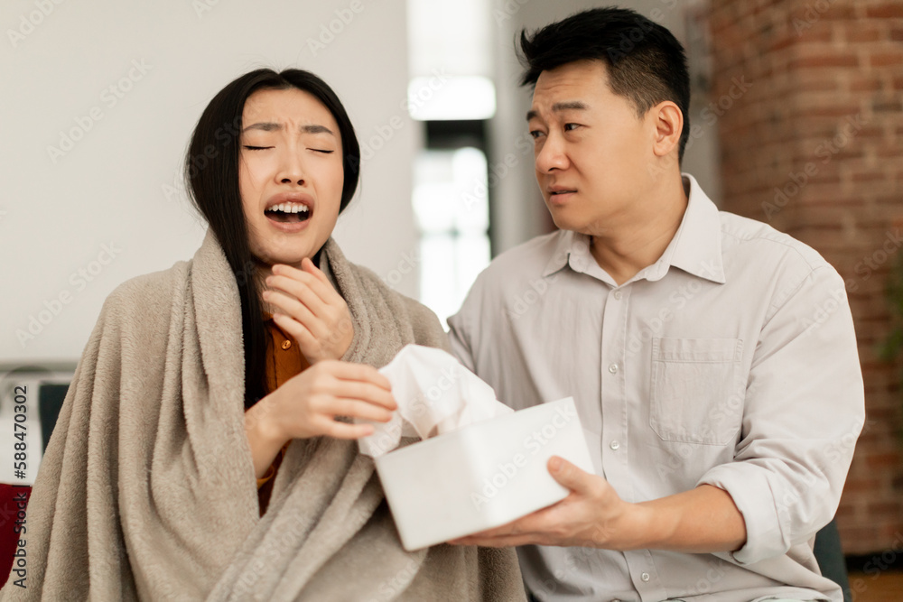 Seasonal allergy and rhinitis illness. Young asian woman sneezing, caring middle aged husband giving napkins to his wife Seasonal allergy and rhinitis illness. Young asian woman sneezing, caring middle aged husband giving napkins to his wife