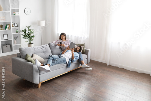 Mixed race spouses resting on sofa at home, using gadgets