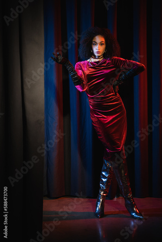 Full length of stylish african american woman in dress and knee boots posing on red background with blue light.