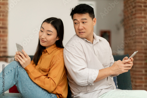 Curious asian middle aged man trying to look at his wife's cellphone, sitting back to back on sofa