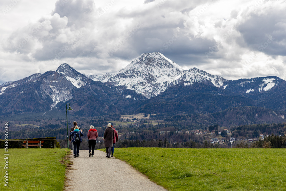 A group of elderly people walking along a village road, between green fields towards the Alps
