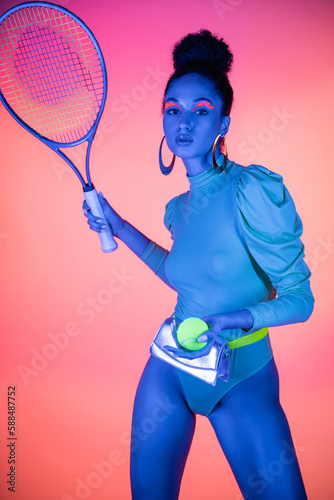 Fashionable african american model holding tennis racket and ball on pink background with neon light. © LIGHTFIELD STUDIOS
