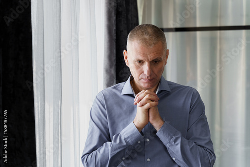 Portrait of a man who stands at the window clenched his hands in front of him, concentrated, prays or thinks