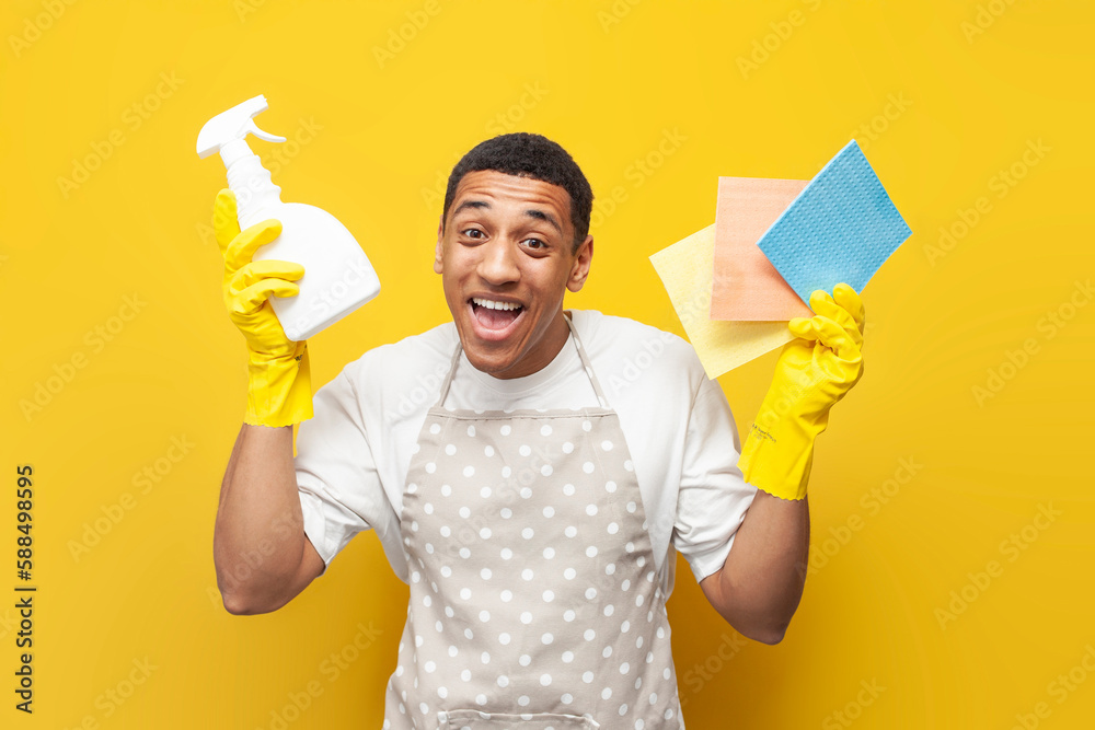 guy african american cleaner in apron and gloves shows rags and detergent on yellow isolated background guy african american cleaner in apron and gloves shows rags and detergent on yellow isolated background