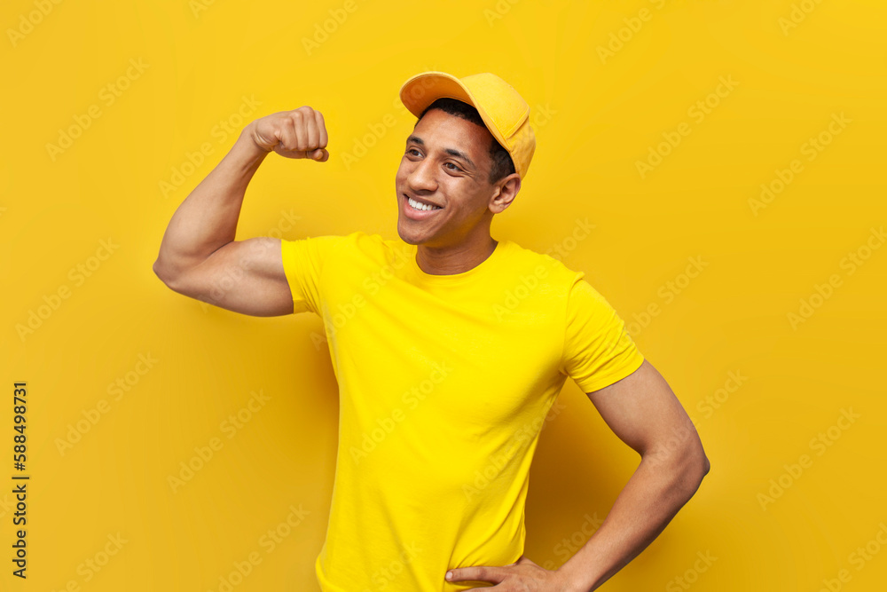 young African American guy in yellow t-shirt and cap shows biceps on yellow background, delivery service worker
