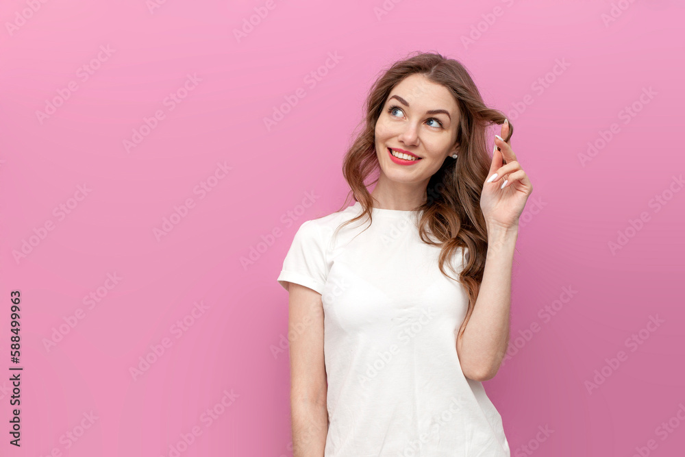 young pensive woman in white t-shirt dreams and thinks on pink isolated background, the girl touches her hair young pensive woman in white t-shirt dreams and thinks on pink isolated background, the girl touches her hair