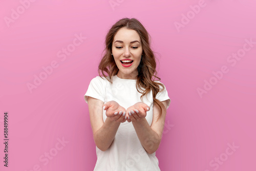 young cute woman in white t-shirt holds empty hands in front of her on pink isolated background