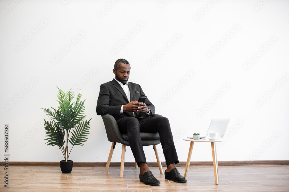 African american businessman in smart suit typing on cellphone while sitting near coffee table with laptop in minimalistic interior. Relaxed manager chatting with clients over internet. African american businessman in smart suit typing on cellphone while sitting near coffee table with laptop in minimalistic interior. Relaxed manager chatting with clients over internet.