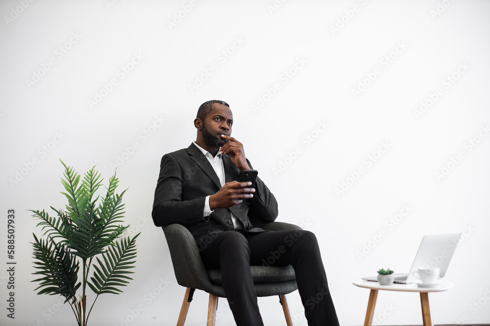 African american man looking aside with pensive facial expression while using modern smartphone at office. Male employer in business suit sitting near coffee table with laptop and cup of drink. African american man looking aside with pensive facial expression while using modern smartphone at office. Male employer in business suit sitting near coffee table with laptop and cup of drink.