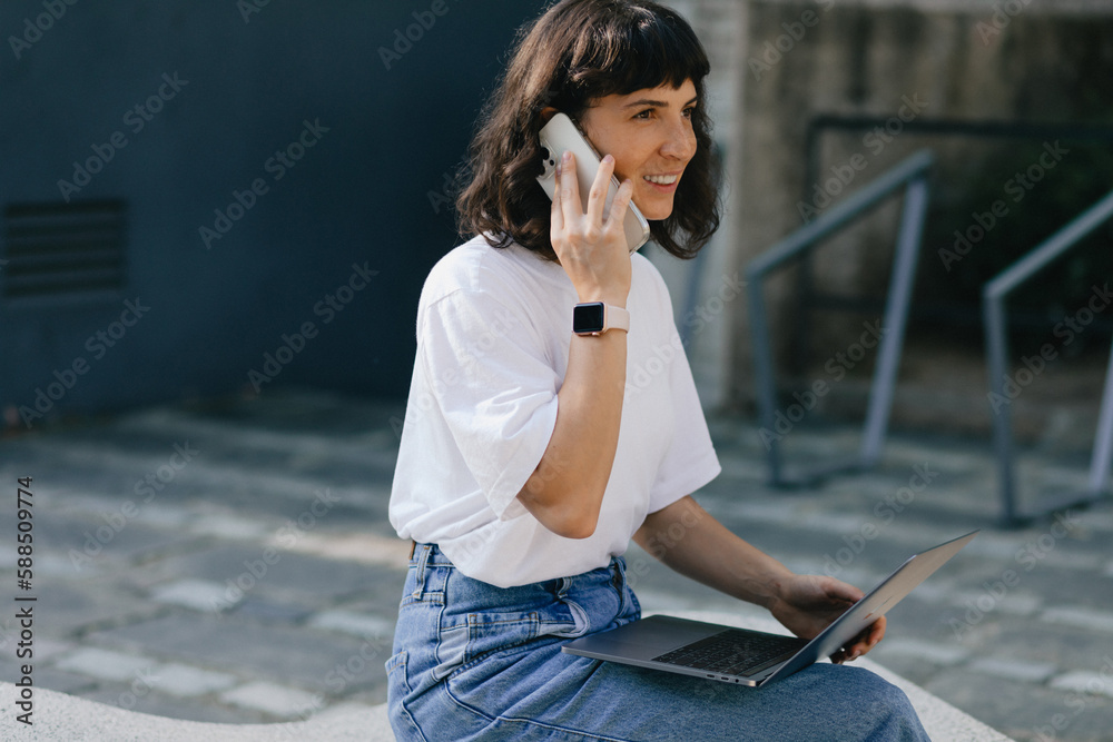 Fototapeta premium Pretty young caucasian girl with gadgets smiling looking aside spending time outdoors. Brunette wears casual clothes. Concept lifestyle, technology in modern society