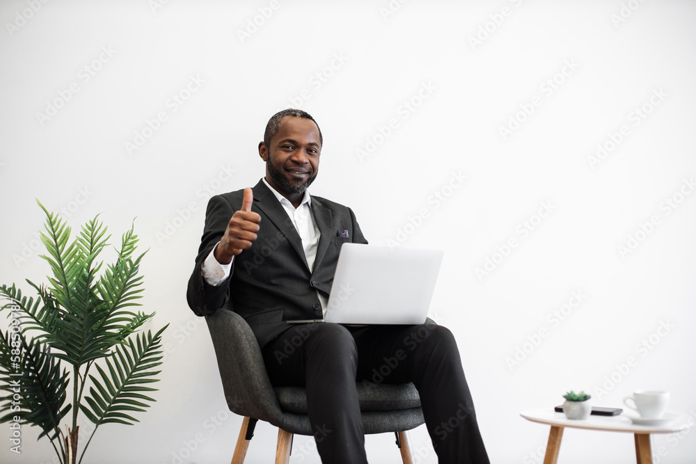 Diligent african employee in formal wear smiling sincerely at camera while typing on wireless laptop at office with modern minimalist design showing thumb up. People, work and technology concept. Diligent african employee in formal wear smiling sincerely at camera while typing on wireless laptop at office with modern minimalist design showing thumb up. People, work and technology concept.