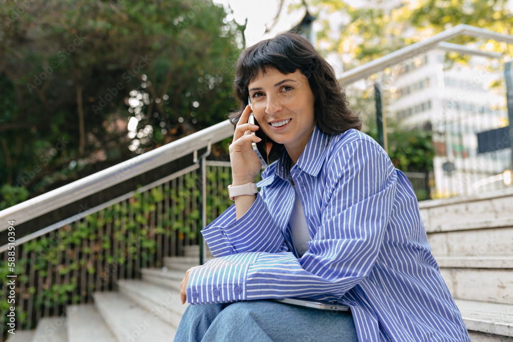 Positive young caucasian woman looking to camera while talking on the phone in city park. Lifestyle concept Positive young caucasian woman looking to camera while talking on the phone in city park. Lifestyle concept