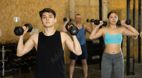 Young dedicated man doing exercises with dumbbells near other people in gym