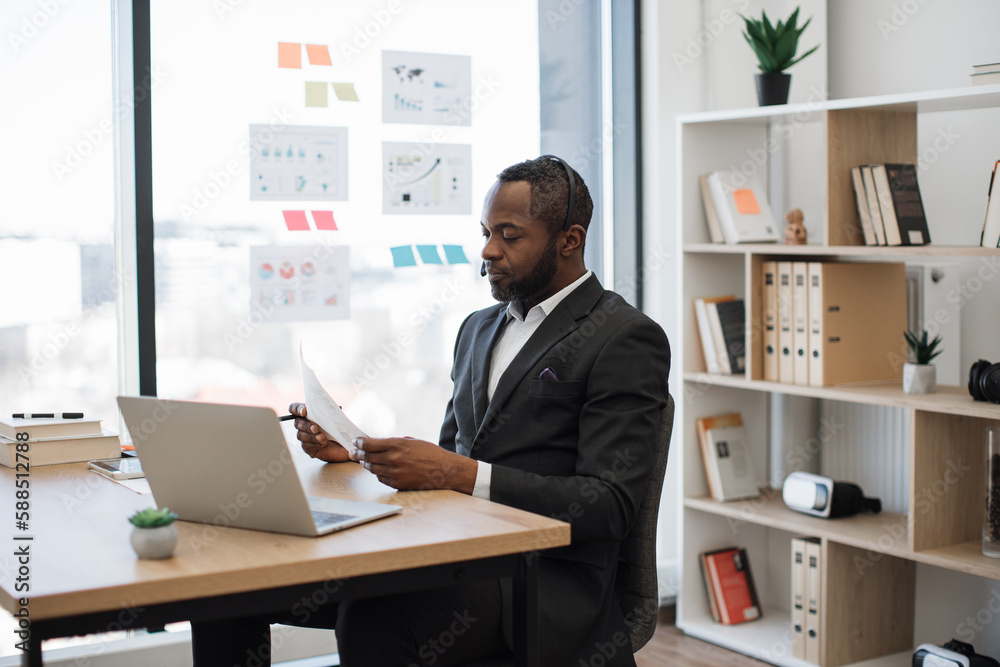 Formally dressed male entrepreneur sitting at workspace and having online meeting with colleagues on laptop. African american man in headset examining some documents holding in hands. Formally dressed male entrepreneur sitting at workspace and having online meeting with colleagues on laptop. African american man in headset examining some documents holding in hands.