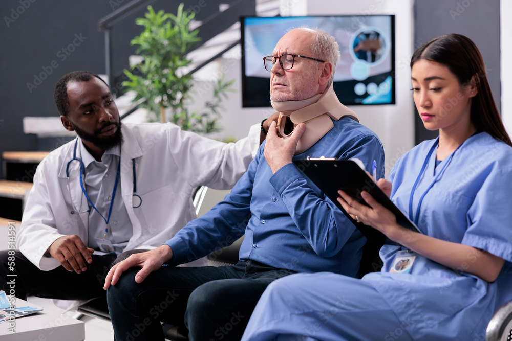 Fototapeta premium Traumatologist doctor explaining rehabilitation therapy to senior patient while assistant typing medical expertise on tablet. Senior man wearing cervical neck collar in hospital waiting area