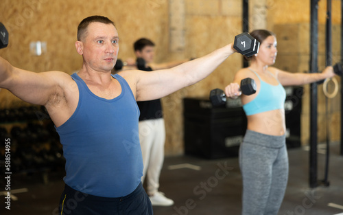 Dedicated active strong fit middle-aged man doing exercises for shoulders while pushing up dumbbells in CrossFit center © JackF